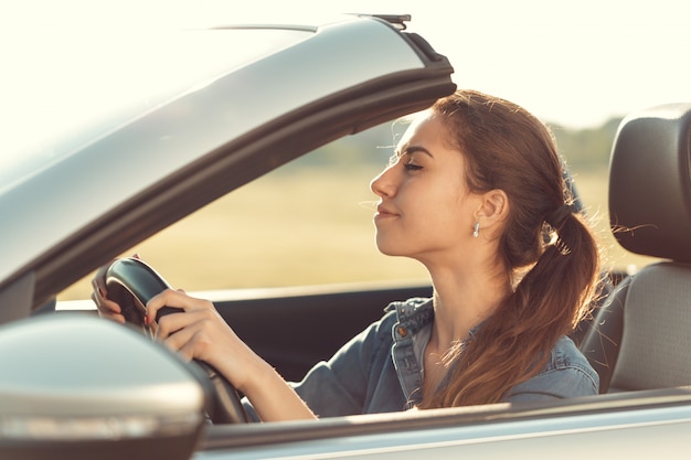 Ragazza che guida l'automobile di cabrio, sulla luce del tramonto