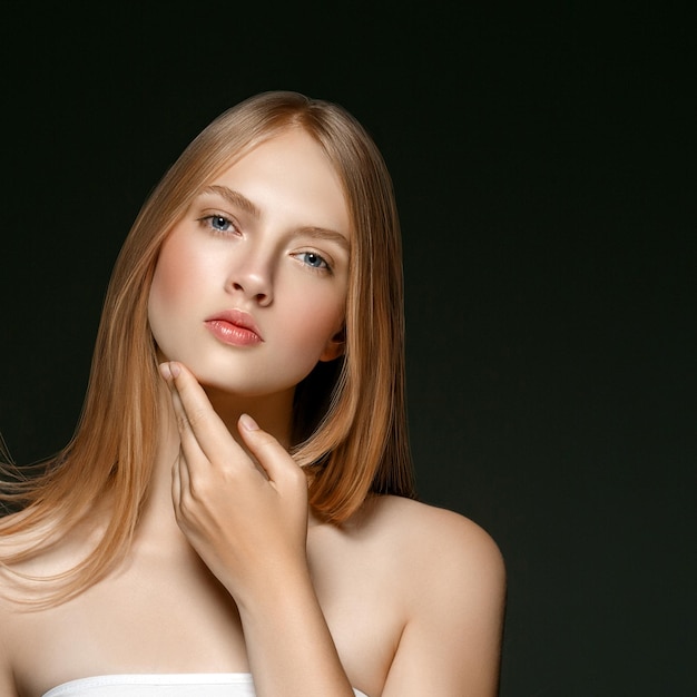 Young girl face beauty skin portrait with long blonde hair with hand over dark background. Studio shot.