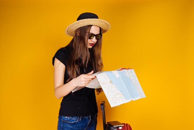 A young girl in a fashionable hat and sunglasses examines a map, goes on a trip with a suitcase