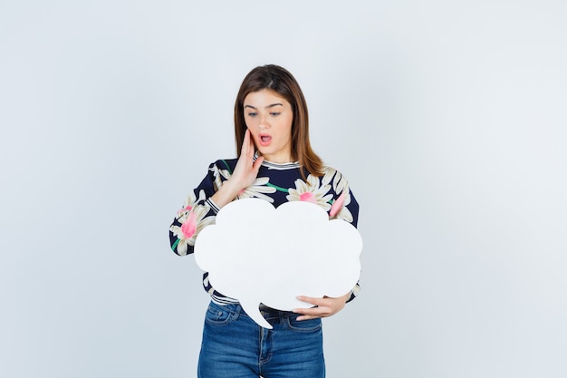 Giovane ragazza in camicetta floreale, jeans che tengono il palmo sulla guancia e sembrano sorpresa, vista frontale.