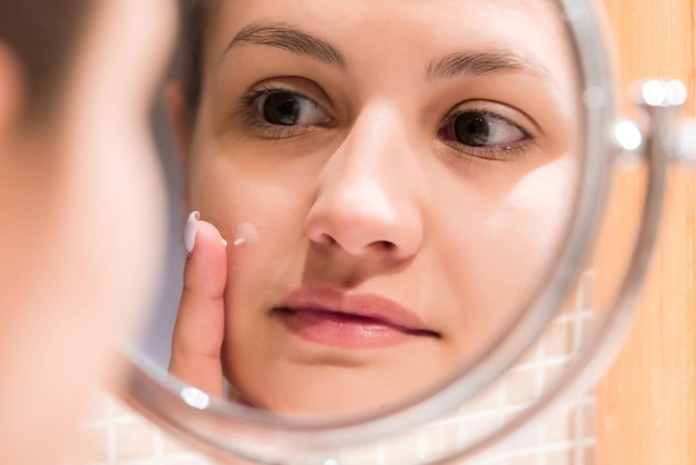 Young girl in front of a bathroom mirror putting cream on a red pimple Beauty skincare and wellness morning concept