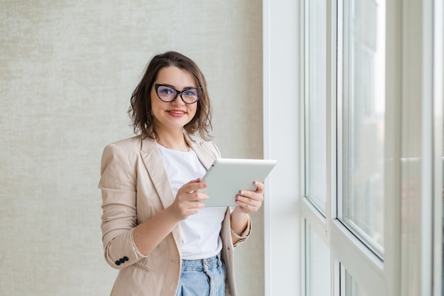 Young girl in glasses for vision stands next to the window of the house holding a tablet