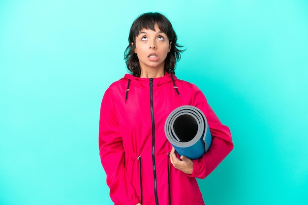 Young girl going to yoga classes while holding a mat isolated on blue background looking up and with surprised expression