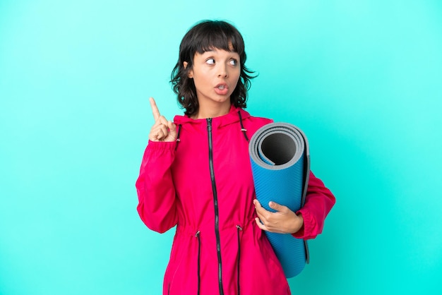 Young girl going to yoga classes while holding a mat isolated on blue background thinking an idea pointing the finger up