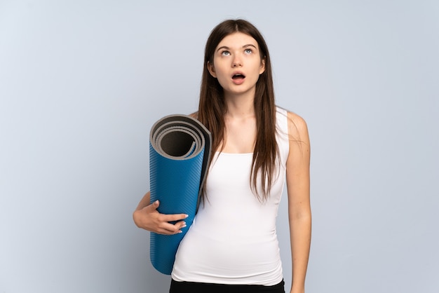 Young girl going to yoga classes while holding a mat looking up and with surprised expression