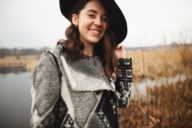 Young girl in gray cardigan and black hat smiles and poses on the shore of a lake