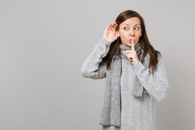 Young girl in gray sweater scarf saying hush be quiet with finger on lips shhh gesture eavesdrop and hearing isolated on grey background. Healthy fashion lifestyle people emotions cold season concept.