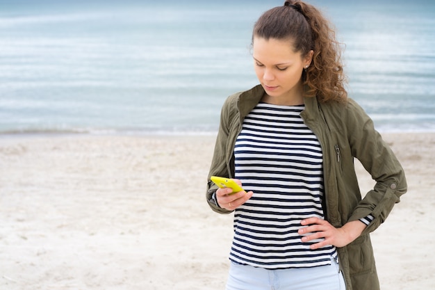 Young girl in a green jacket uses yellow mobile phone on the beach