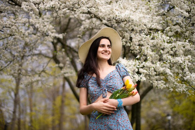 A young girl in a hat and with tulips standing in a blue dress next to a blooming white tree