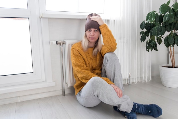 A young girl in a hat and a yellow sweater is sitting on the floor and holding her head near a heater with a thermostat