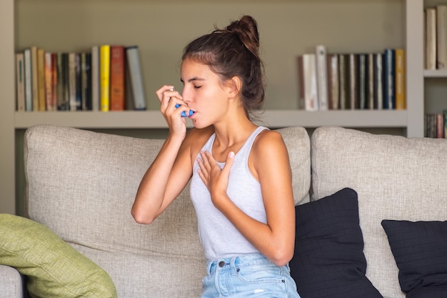 Молодая девушка с приступом астмы и с помощью ингалятора, сидя на диване дома