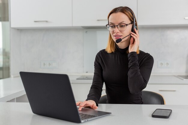 A young girl in a headset with a microphone works in a call center sitting at a laptop at home in the kitchen. High quality photo