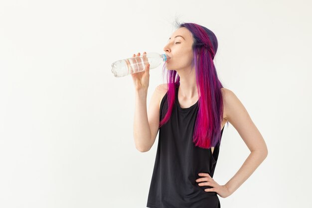 Young girl hipster with colored hair drinks water from a bottle after intense physical training in the gym. The concept of thirst and water balance of the body. White background with copy space