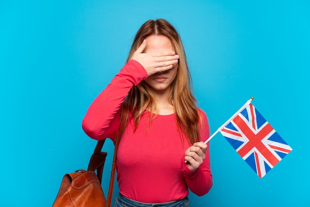 Фото Молодая девушка держит флаг соединенного королевства на изолированном синем фоне, закрывая глаза руками. не хочу что-то видеть