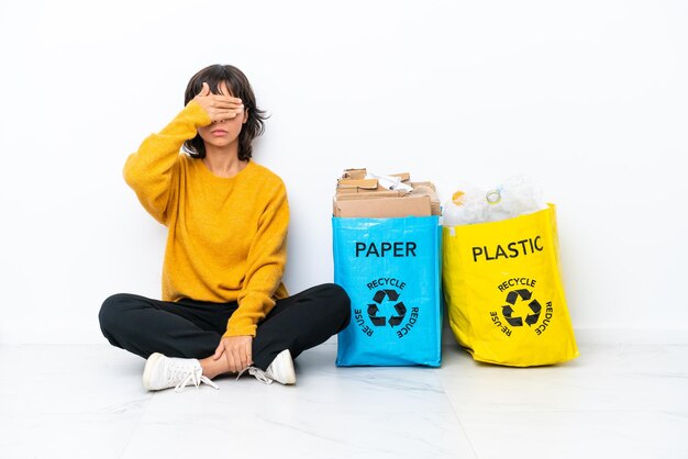 手で目を覆っている白い背景で隔離の床に座ってプラスチックと紙でいっぱいのバッグを保持している少女。何かを見たくない