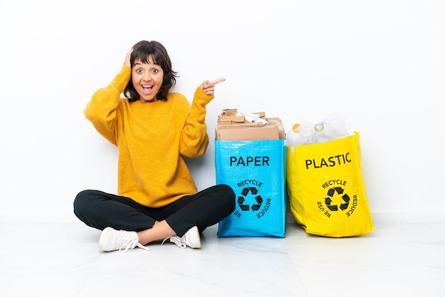 白い背景で隔離の床に座ってプラスチックと紙でいっぱいのバッグを持っている少女は驚いて、人差し指を横に向ける