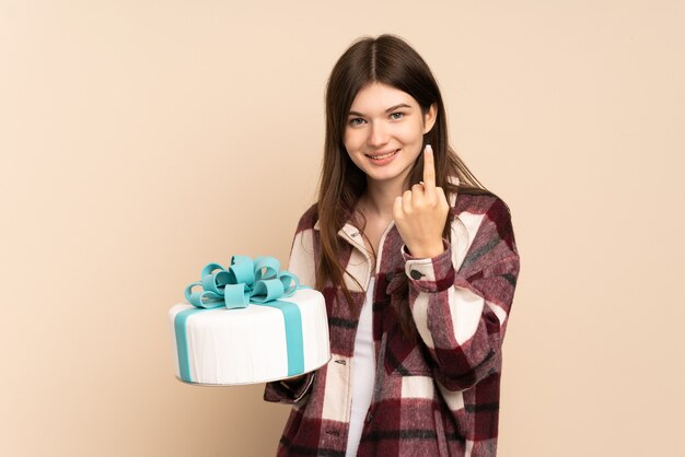 Young girl holding a big cake isolated on beige doing coming gesture