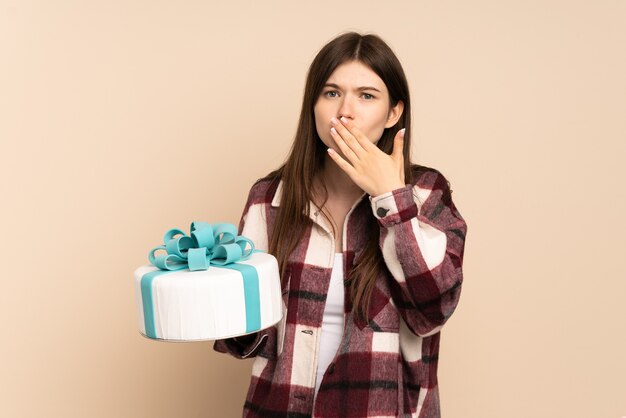 Young girl holding a big cake isolated on beige happy and smiling covering mouth with hand