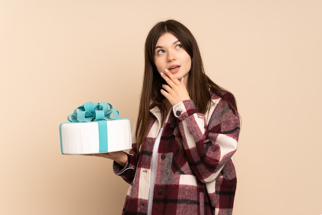 Young girl holding a big cake isolated on beige looking up while smiling