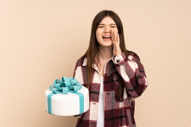 Young girl holding a big cake isolated on beige shouting with mouth wide open