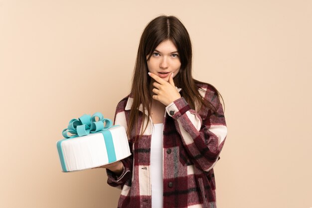 Young girl holding a big cake isolated on beige thinking