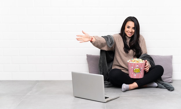 Young girl holding a bowl of popcorns and showing a film in a laptop presenting and inviting to come with hand