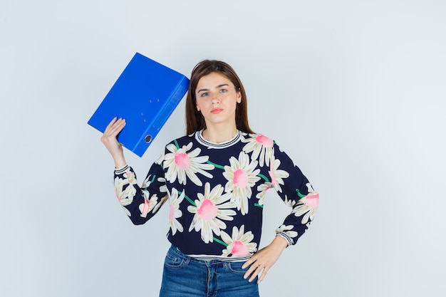 Photo young girl holding folder near head in floral blouse, jeans and looking puzzled , front view.