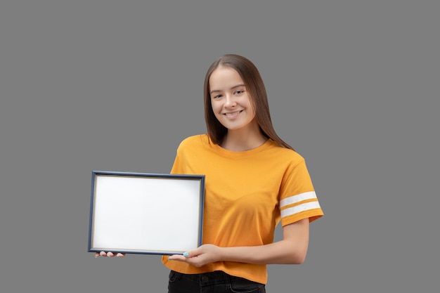 Young girl holding a horizontal blue frame with white mockup in her hands smiling isolated on a gray background