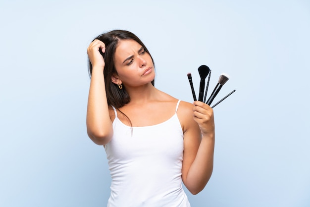 Young girl holding a lot of makeup brush having doubts and with confuse face expression