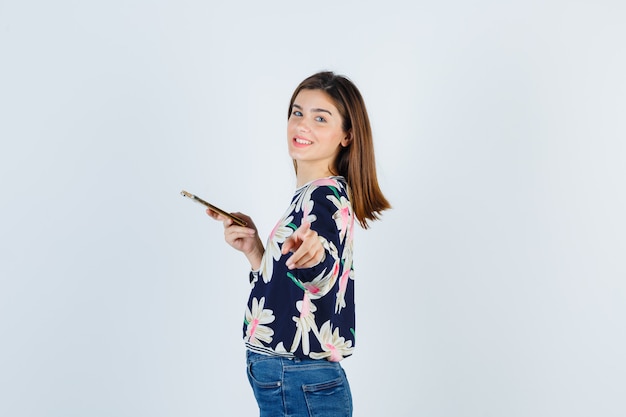 Молодая девушка держит телефон, указывая на камеру, боком стоит в цветочной блузке, джинсах и выглядит веселой.