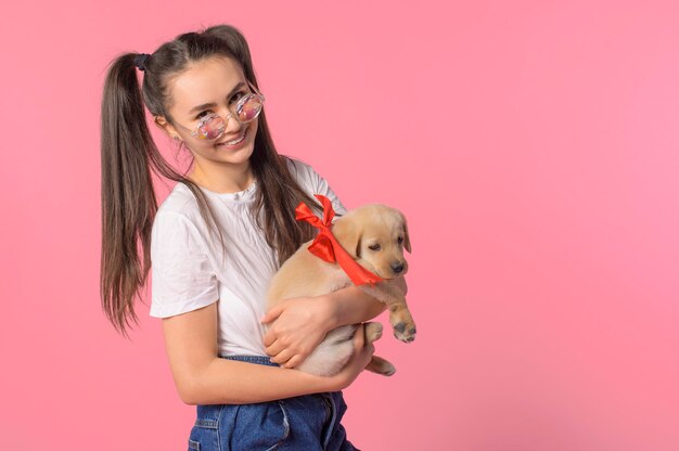 Foto la ragazza che tiene un cucciolo con un nastro rosso sulle braccia sorride e felice su uno sfondo rosa