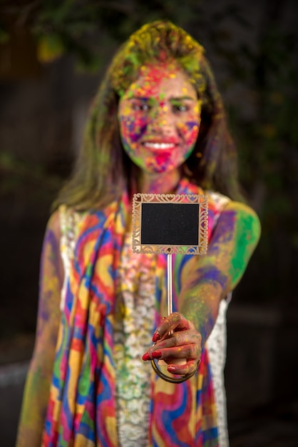 컬러 스플래시와 함께 가루 색상으로 칠해진 얼굴로 Holi 축제 행사에 작은 보드를 들고 어린 소녀.