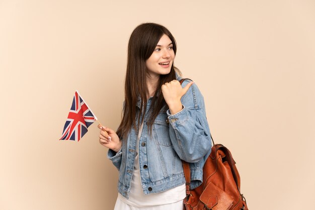 Young girl holding an United Kingdom flag isolated on beige pointing to the side to present a product