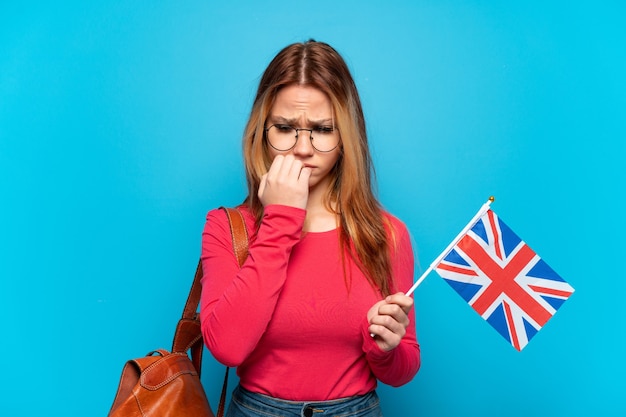 Young girl holding an United Kingdom flag over isolated blue background having doubts