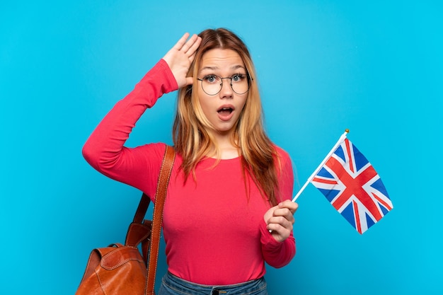 Young girl holding an United Kingdom flag over isolated blue wall with surprise expression