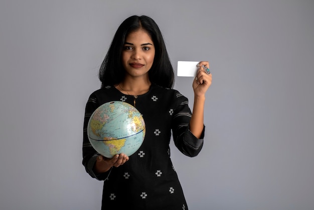 Young girl holding the world globe and posing with credit card on a grey wall.