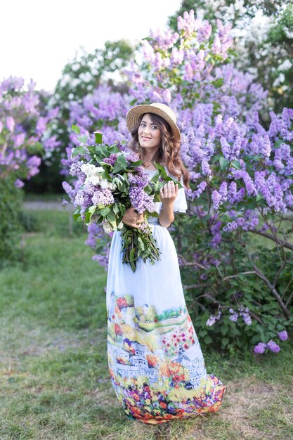 若い女の子は花束ライラックの花を保持します夏の庭に立っている麦わら帽子の女の子