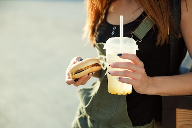 A young girl holds a drink and a cheeseburger in the hand against the background of a sunny street