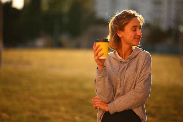 若い女の子は彼の手にコーヒーのグラスを持っています。女の子は通りの紙コップからlatを飲みます