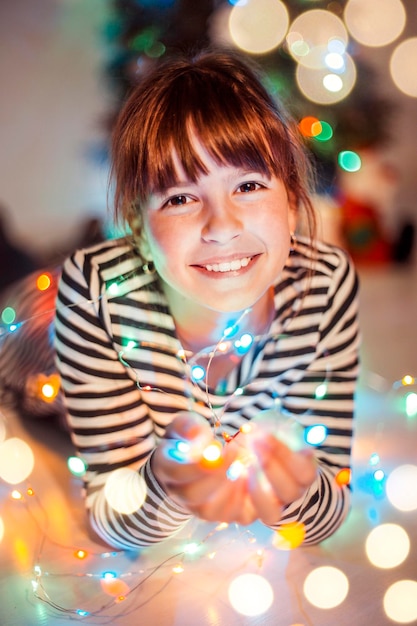 Young girl holds light colorful garland in hands before Christmas Tree