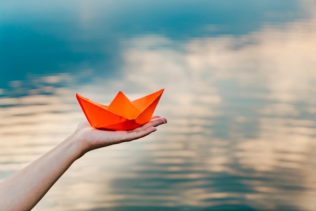 A young girl holds a paper boat in her hand above the river. Origami in the form of a ship has an orange color