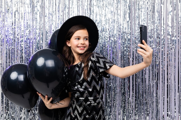 Young girl on holiday takes a photo on a smartphone among black balloons on a shiny