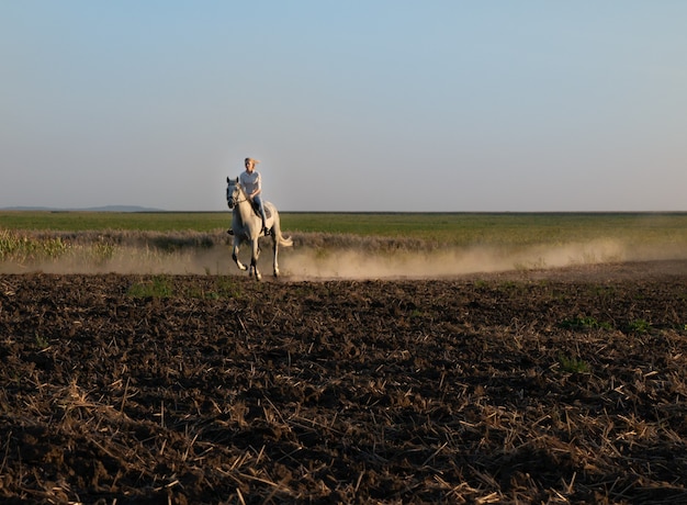 A young girl on horseback rides across the field during sunset.