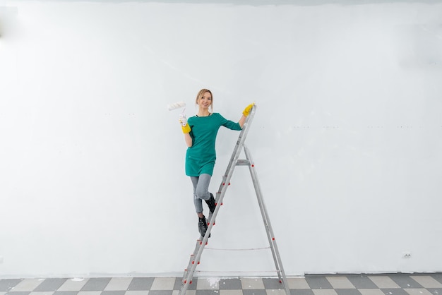 A young girl is engaged in repairs on the stairs and paints a\
white wall with a roller in a new apartment renovation of the\
interior and a new apartment housewarming and a desirable\
mortgage