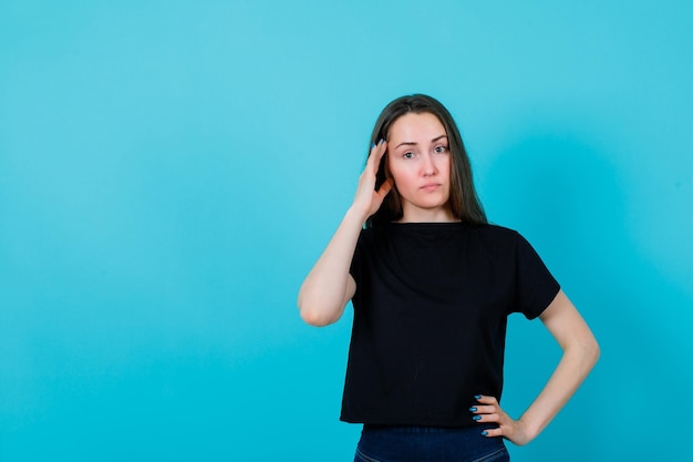 Young girl is holding hand on temple and putting other on waist on blue background