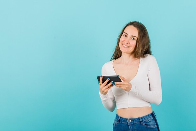 Young girl is looking at camera by holding smartphone on blue background