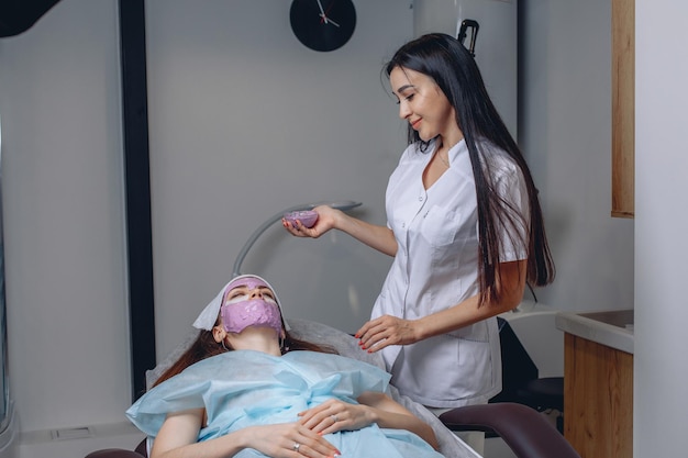 A young girl is lying on a couch in a beauty salon with her face completely covered with a mask for self-care. There is a cosmetologist nearby.