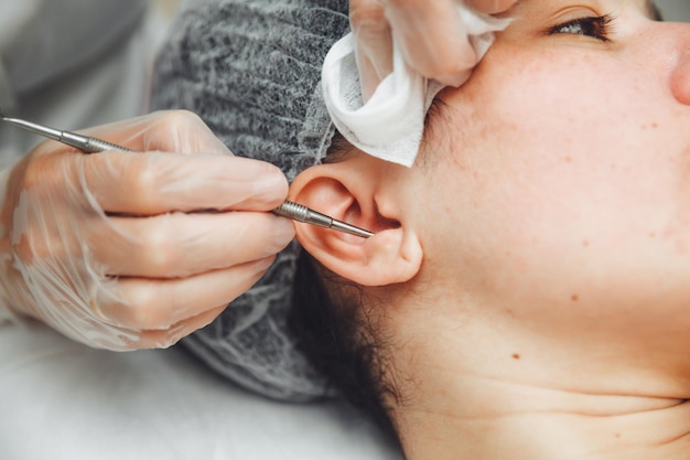 A young girl is lying on a couch during cosmetic procedures\
over which a cosmetologist squeezes out fat deposits and pimples\
with a special metal toolear cleaning