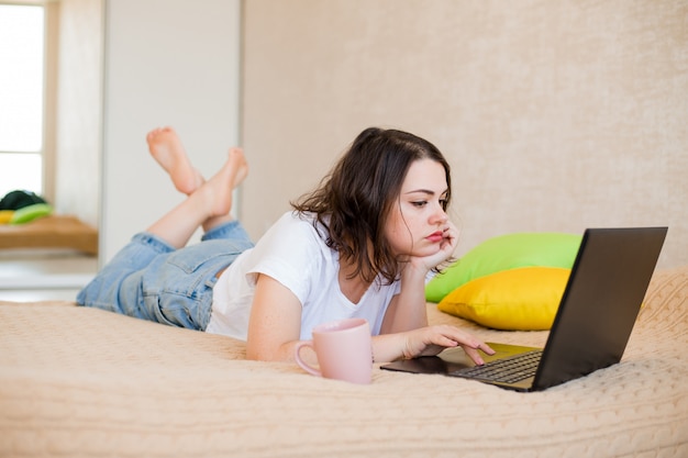 Young girl is lying at home on a bed with coffee and a laptop