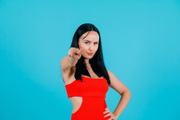 Young girl is pointing to camera with forefinger and putting hand on waist on blue background